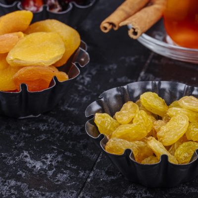 side view of dried raisins and dried apricots in mini tart tins served with tea on black wooden background side view of dried raisins and dried apricots in mini tart tins served with tea on black wooden background